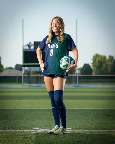 Allie Louvier #8 full length on the field, holding the ball on her hip and smiling