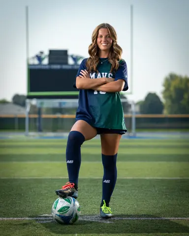 Kora De Burgh #18 full length, arms crossed with one foot on the ball, scoreboard behind her
