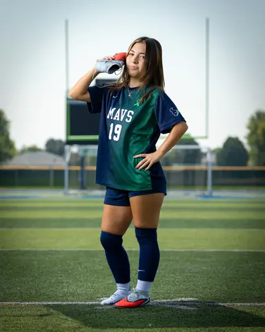 Maria Rojas #19 full length, holding a cleat to her ear like a phone, hand on hip