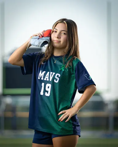 Maria Rojas #19 waist up, holding a cleat to her ear like a phone, hand on hip