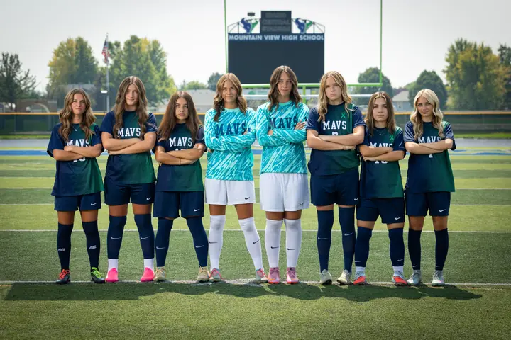 The eight Mountain View girls soccer seniors lined up full length with arms crossed in front of the scoreboard