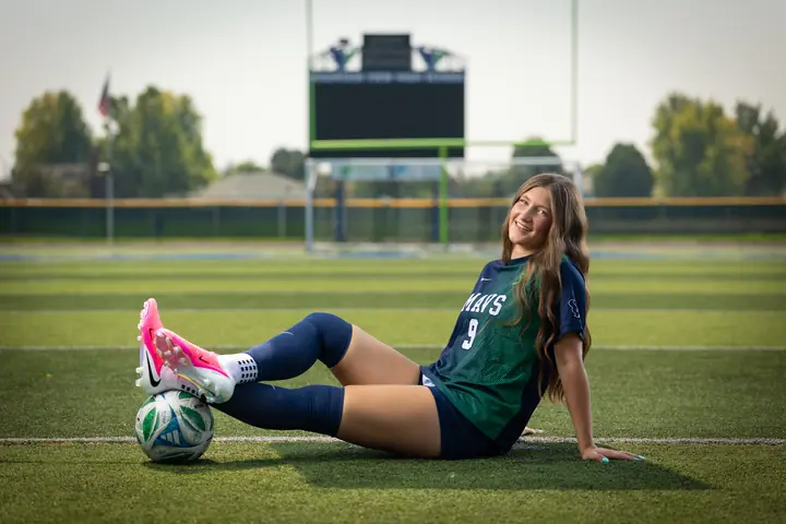 Vivian Sipe #9 sitting on the turf, smiling, pink cleats resting on a soccer ball