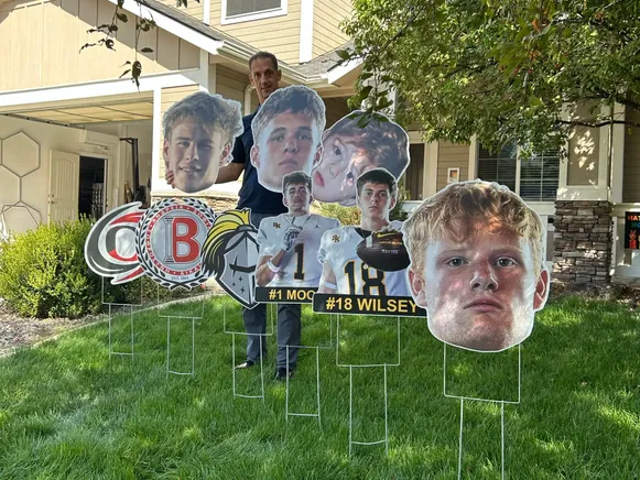 Mark Rezansoff of Shooting Trio on a front lawn full of big heads, school logo cutouts, and player signs