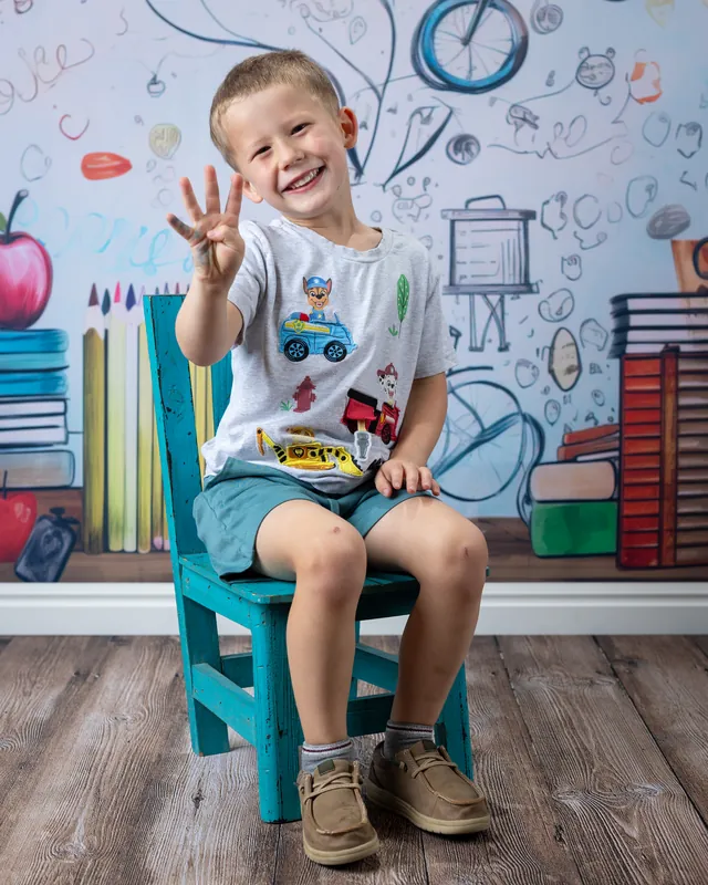 Smiling boy waving from a little blue chair in front of a school doodle backdrop at daycare photo day - preschool photography Meridian Idaho