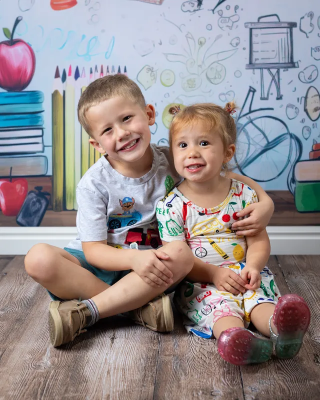 Brother and little sister sitting together on the floor in front of a school doodle backdrop at daycare photo day - sibling portraits Meridian Idaho
