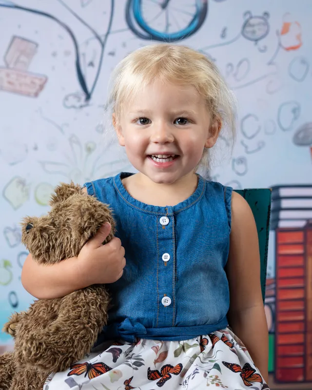 Toddler hugging a teddy bear on a little chair with a school doodle backdrop at daycare picture day - preschool photography Meridian Idaho