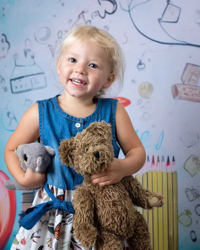 Smiling toddler holding a teddy bear in front of a hand-drawn school doodle backdrop at daycare photo day - daycare photographer Meridian Idaho