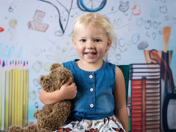 Toddler hugging a teddy bear in front of a school-themed backdrop at a Meridian daycare photo day